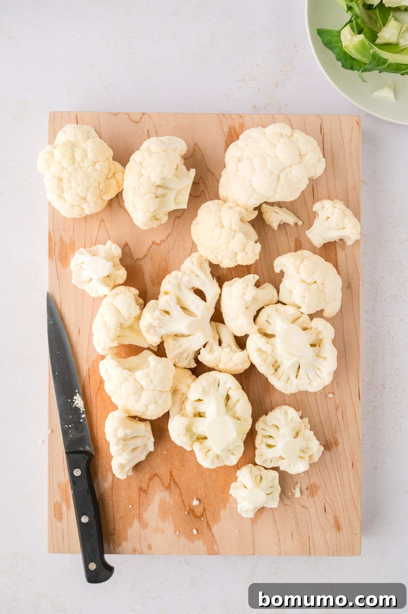 A chef's knife carefully slicing a head of cauliflower into individual florets on a cutting board, emphasizing preparation.