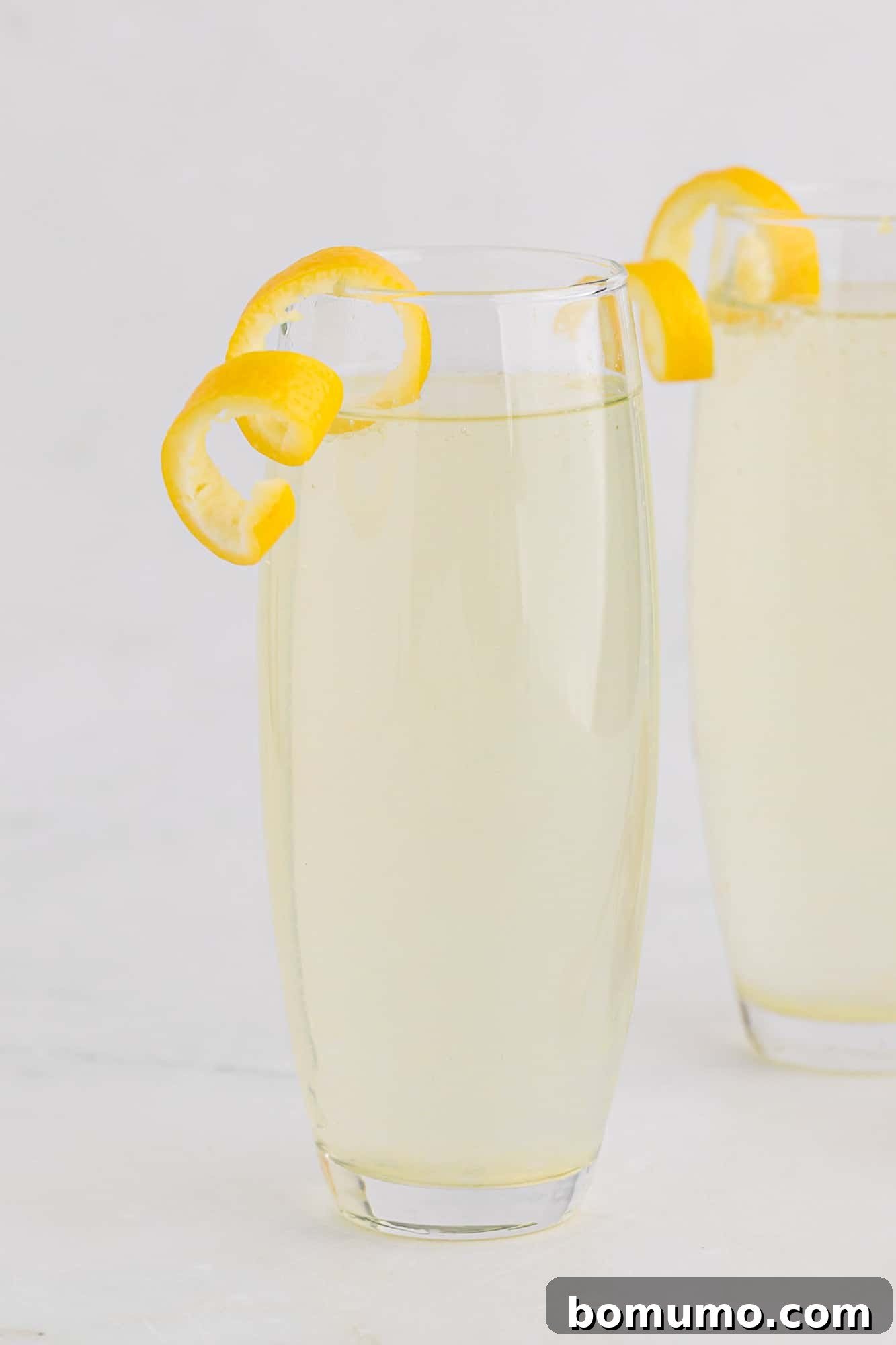 Two elegant French 75 cocktails, one in the foreground with a vibrant lemon twist garnish, and another softly blurred in the background, set against a bright, inviting backdrop, emphasizing the cocktail's refreshing and sophisticated appeal.