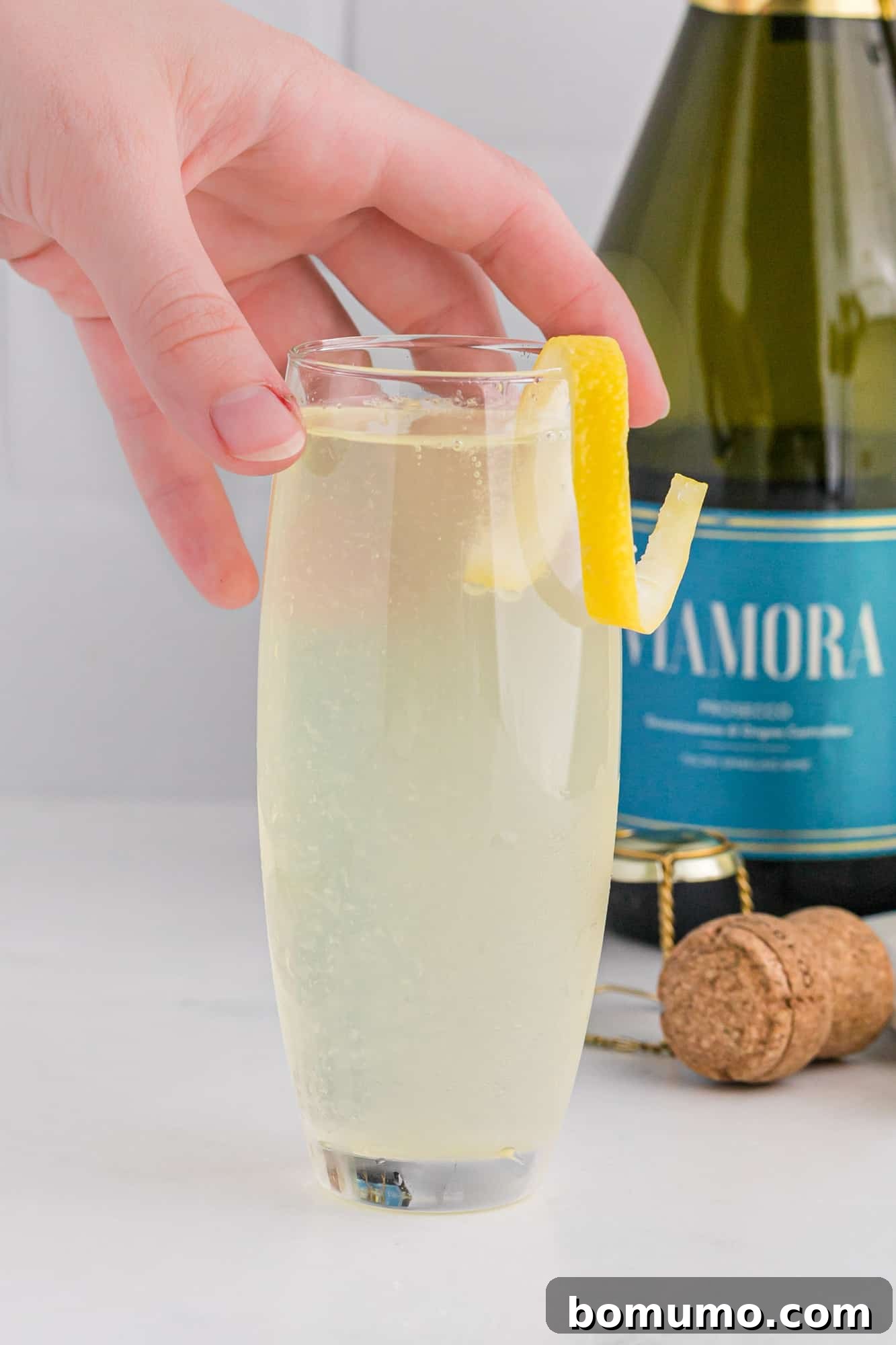 A hand gracefully holds a stemless champagne flute containing a beautifully garnished French 75 cocktail, inviting one to experience its refreshing taste and elegant presentation.