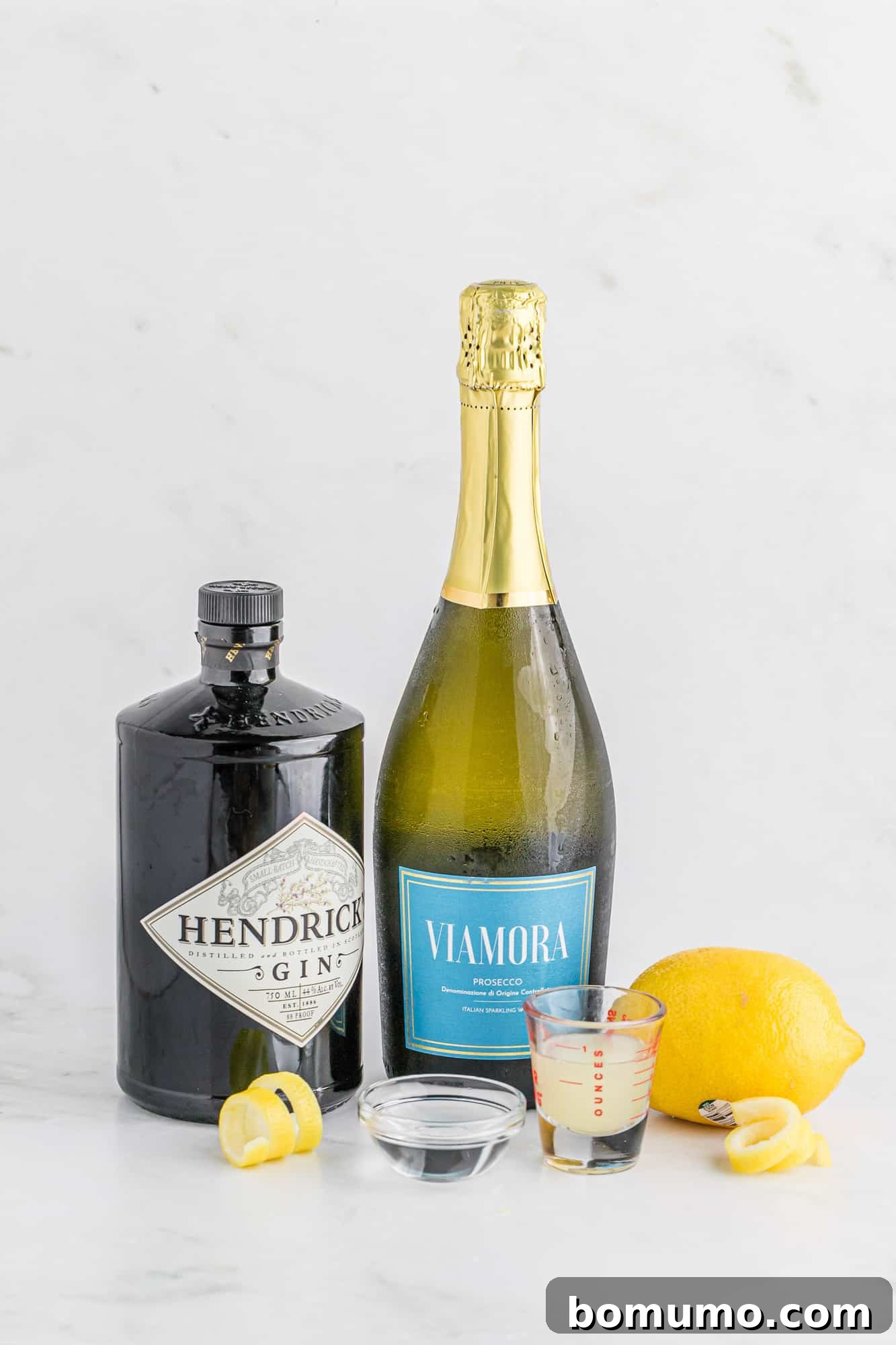 Key ingredients for a French 75 cocktail laid out: a bottle of gin, a bottle of prosecco or sparkling wine, fresh lemons, and a bottle of simple syrup, indicating preparedness for cocktail creation.