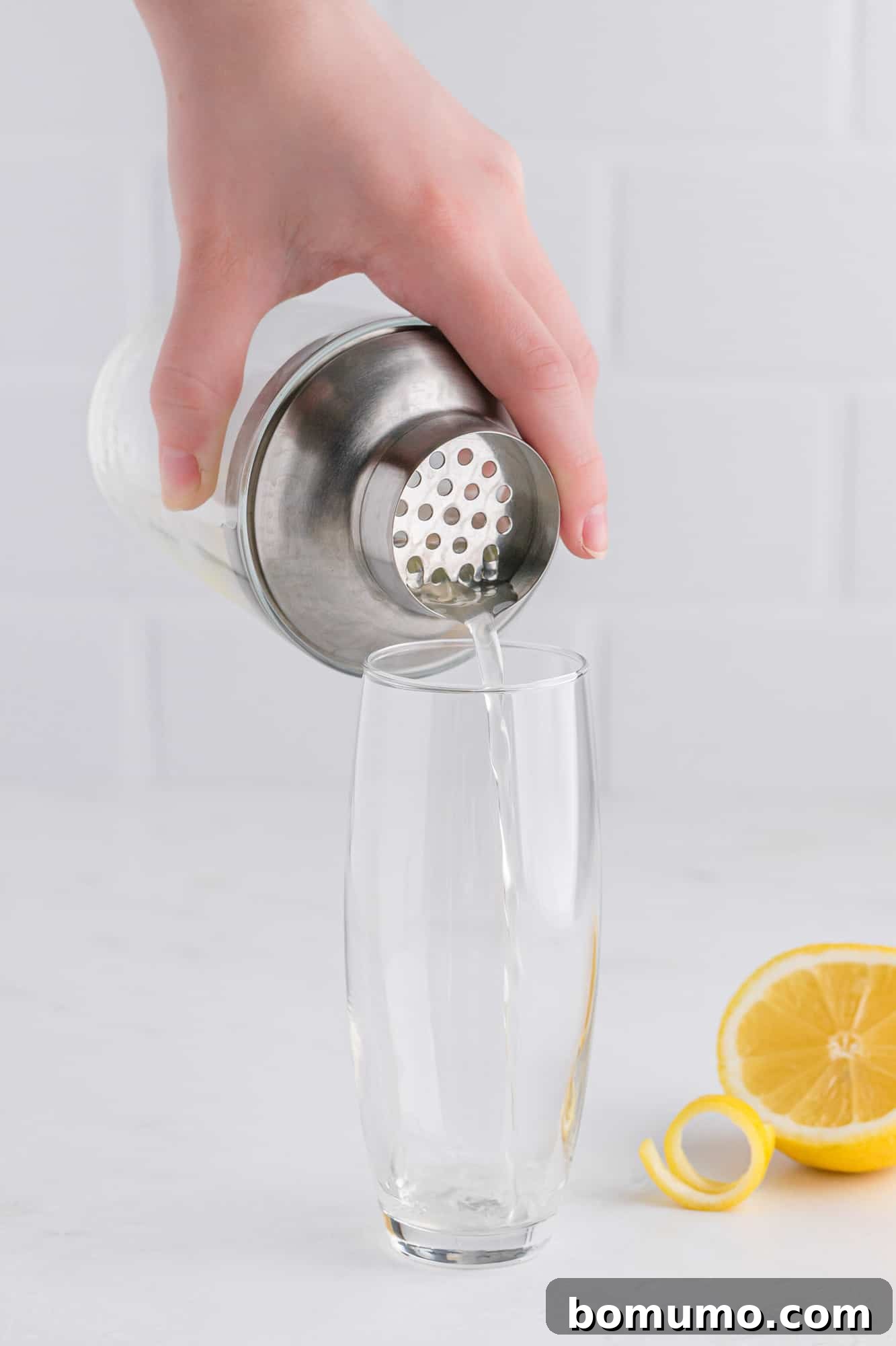A champagne flute being filled with the shaken gin, lemon juice, and simple syrup mixture from a cocktail shaker, illustrating the careful pouring stage of preparing a French 75.