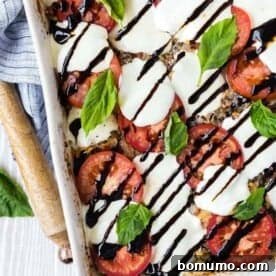 An overhead image of a vibrant Caprese quinoa bake, generously sprinkled with fresh basil and elegantly drizzled with a rich balsamic reduction, presented in a white baking dish next to a wooden spoon and linen.