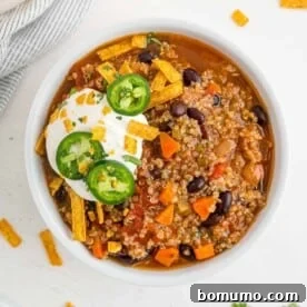 A round bowl of vibrant quinoa chili with black beans, generously garnished with sour cream and sliced jalapeños.