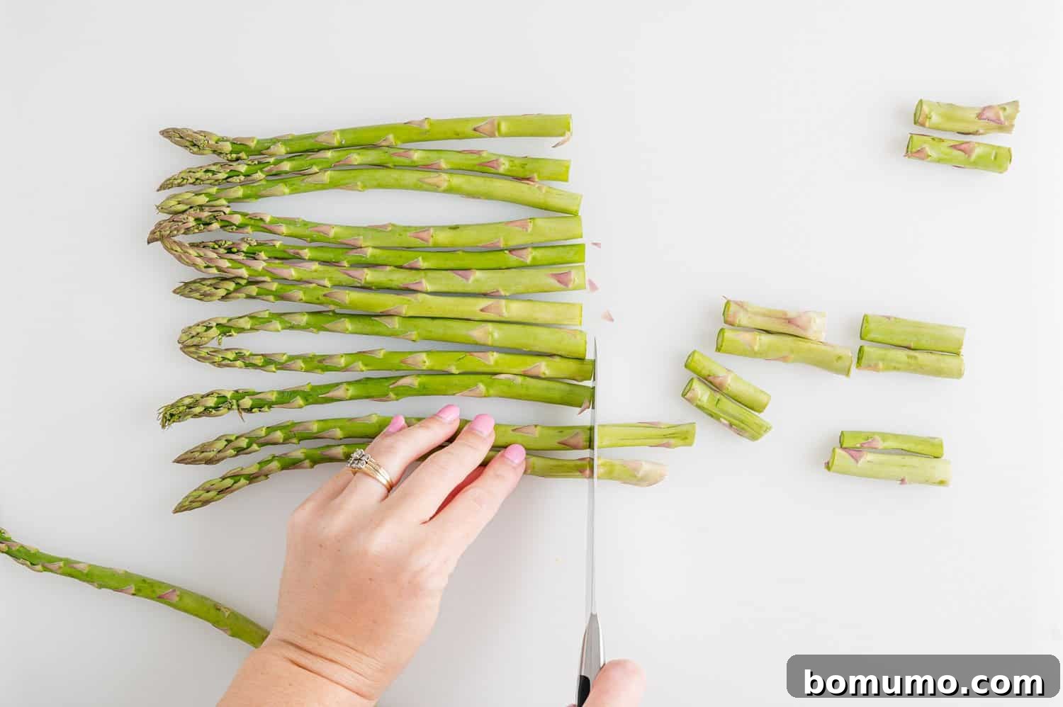 A hand demonstrating how to cut the woody ends off asparagus spears with a sharp knife, ensuring uniform length for cooking.