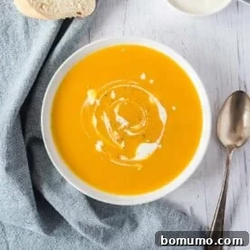 Overhead view of a bowl of pumpkin soup swirled with cream, next to bread slices and a spoon.