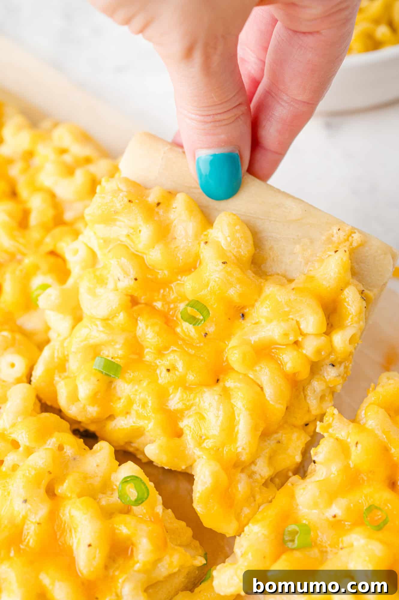 Ultimate Comfort Pizza 13 A hand holds a single, perfectly golden slice of mac and cheese pizza, showcasing the thick, creamy topping and crispy crust.