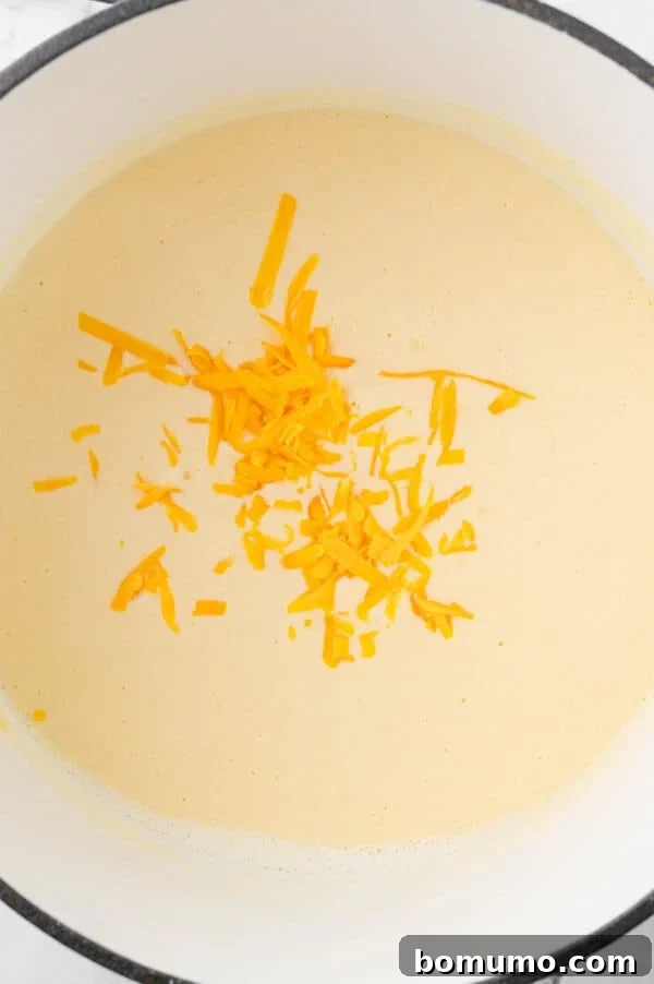 Ultimate Comfort Pizza 6 Shredded cheddar cheese being added to a bubbling white sauce, beginning to melt and create a creamy texture.