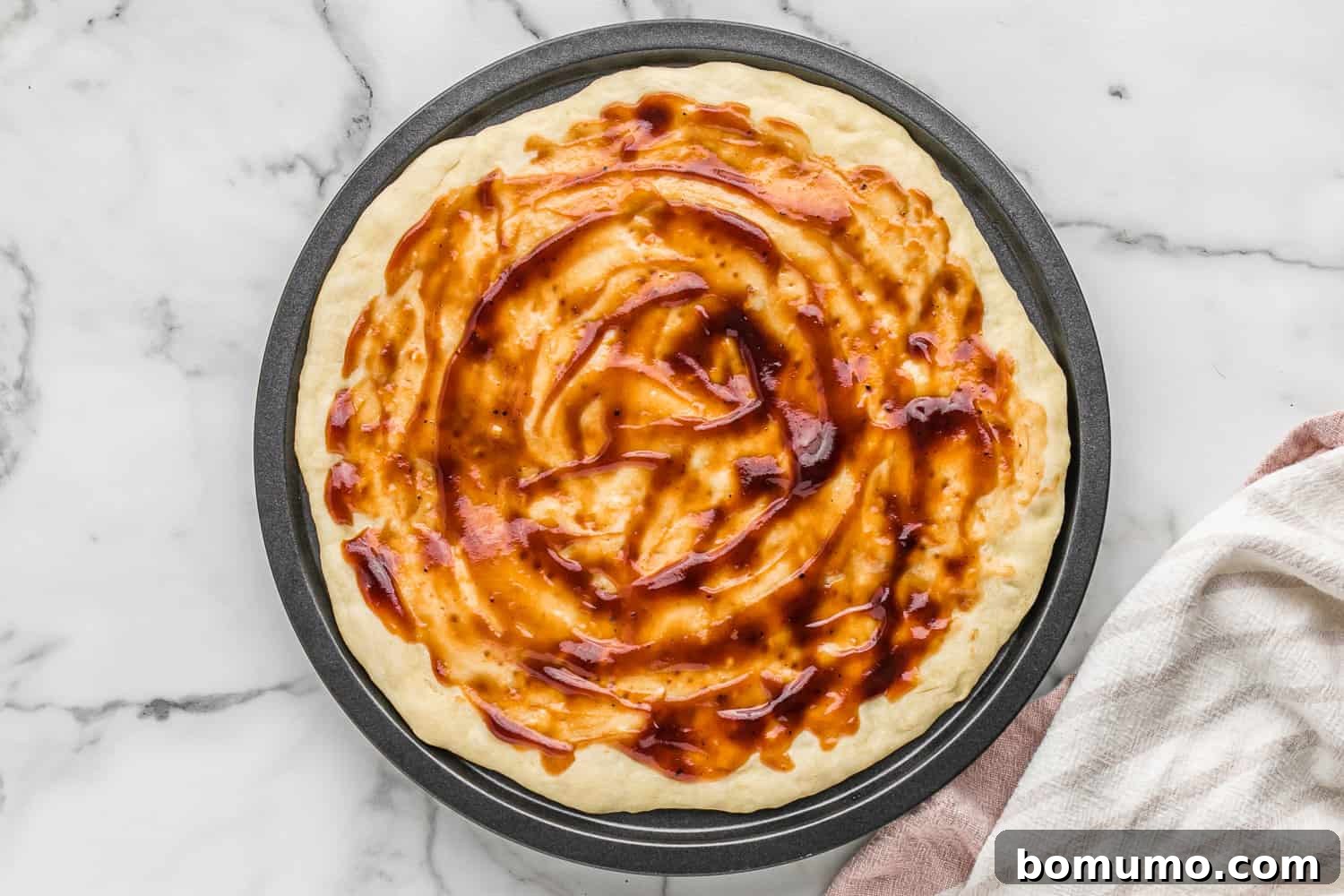 An unbaked pizza crust generously brushed with zesty BBQ sauce, creating a vibrant red base for upcoming toppings.