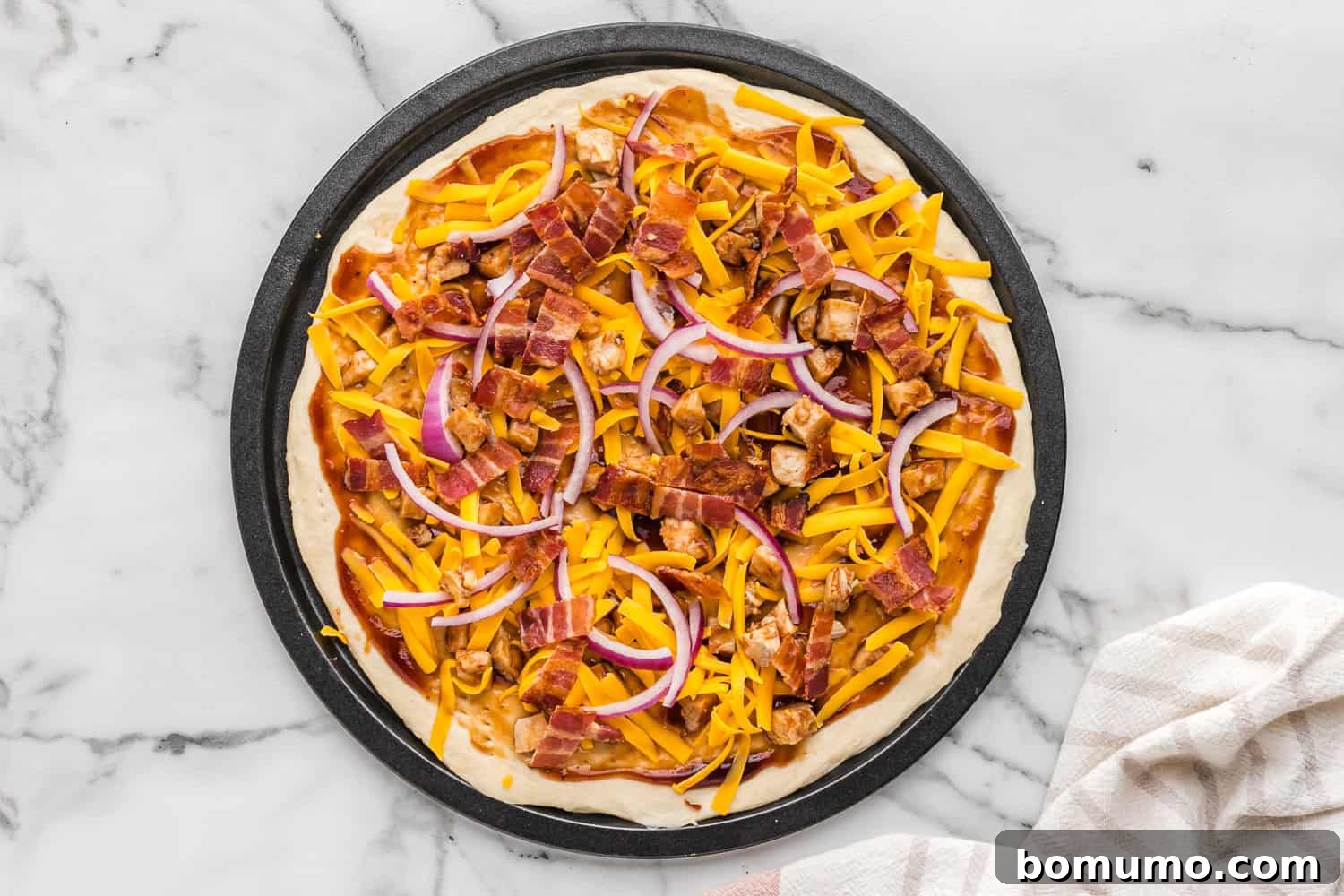 A fully assembled, unbaked BBQ chicken pizza, loaded with chicken, bacon, onions, and cheddar cheese, ready for the oven.