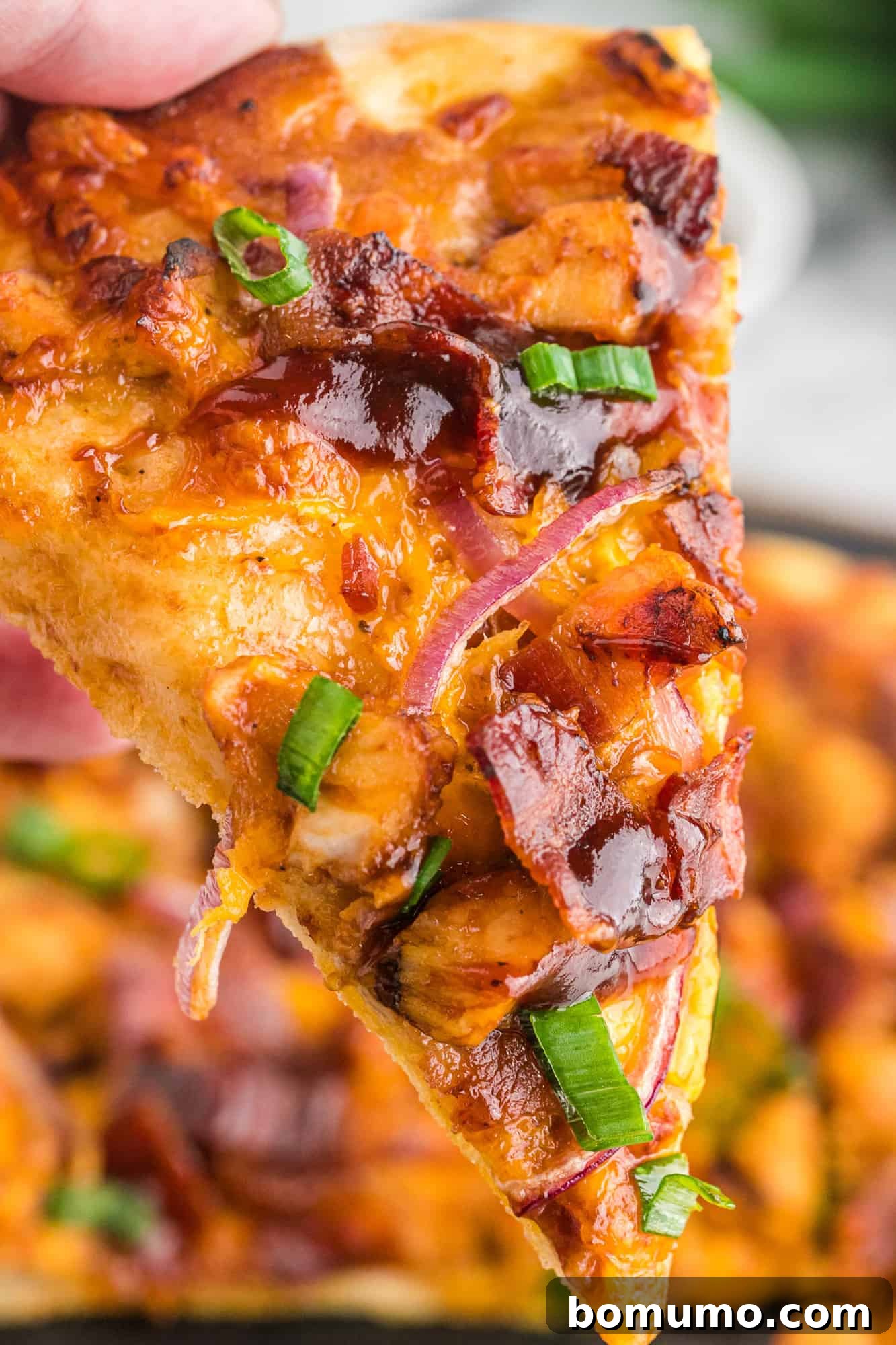 A hand holding a perfectly triangular slice of hot, fresh BBQ chicken pizza, highlighting its appealing crust and ample toppings.