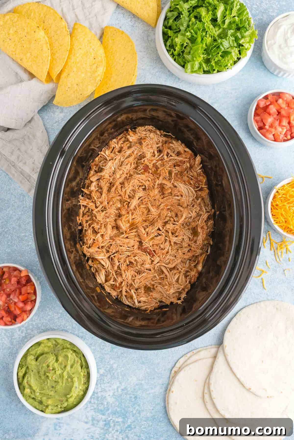 Taco chicken in a slow cooker, surrounded with toppings.