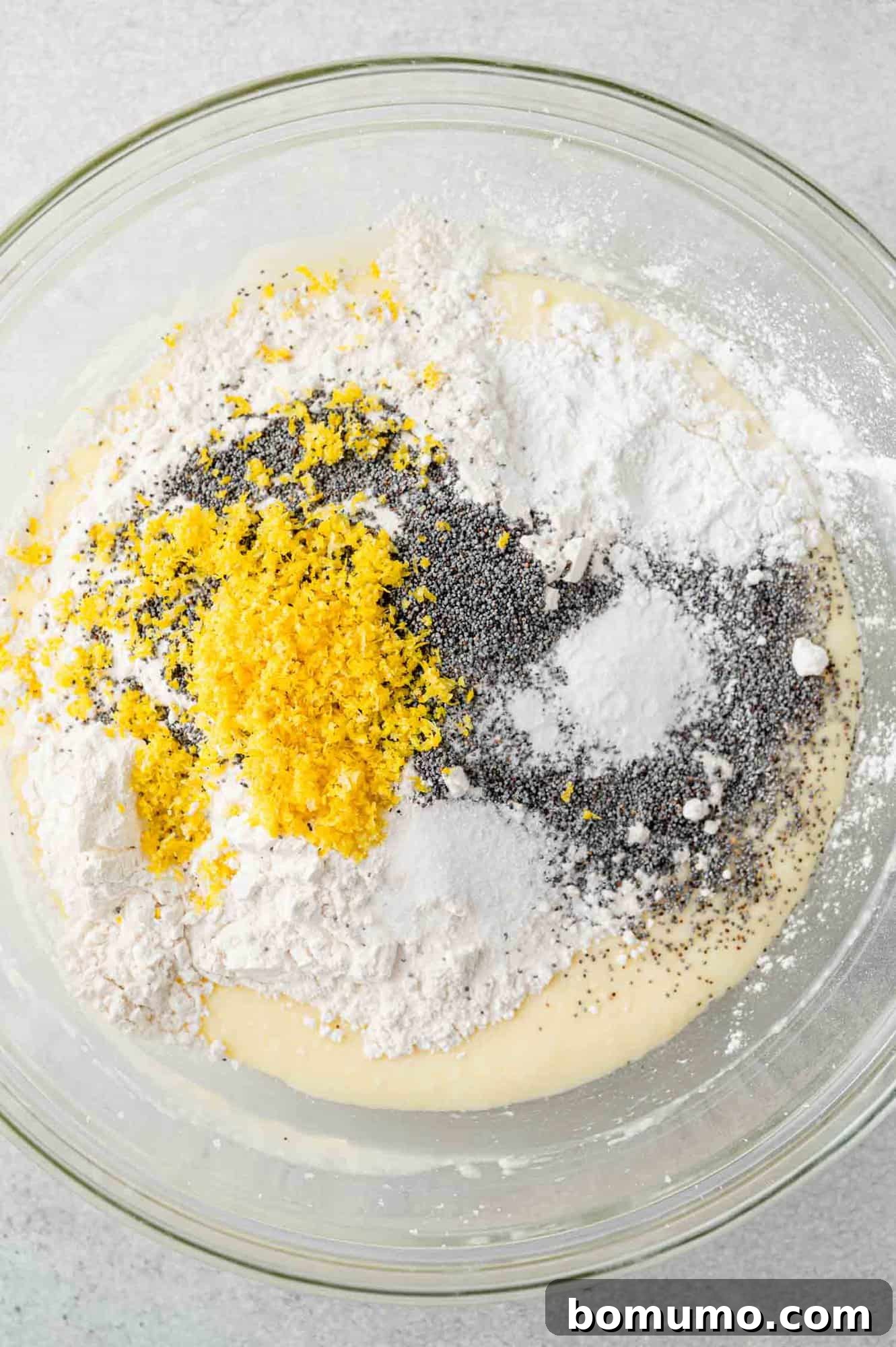 Dry ingredients including flour, poppy seeds, and lemon zest, piled on top of the wet batter in a mixing bowl, prior to being mixed.