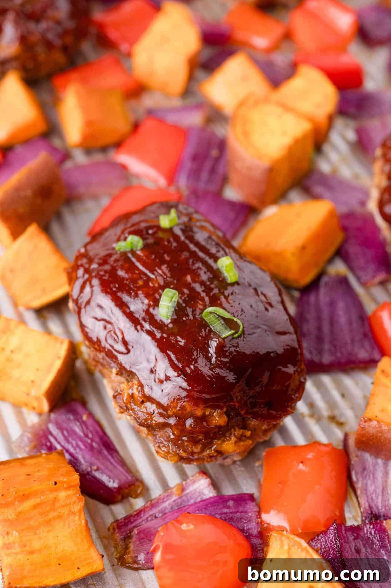 Sheet Pan BBQ Mini Meatloaves 2 Mini BBQ-glazed meatloaf topped with chopped green onions is the star of this easy mini meatloaf recipe, surrounded by roasted sweet potato cubes, red bell peppers, and red onion pieces on a baking sheet.