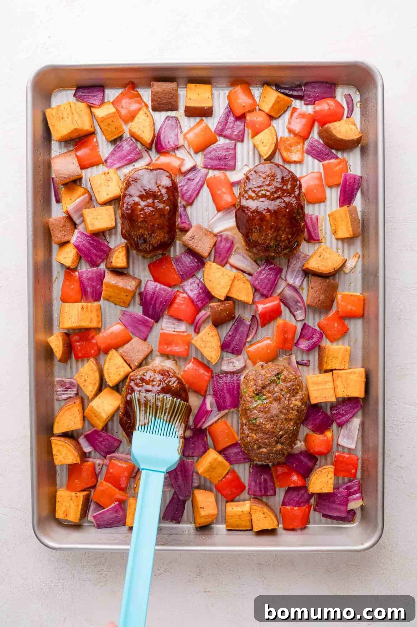 Sheet Pan BBQ Mini Meatloaves 11 A baking sheet with four mini meatloaves, chopped sweet potatoes, red onion, and red bell pepper; a hand brushes sauce onto a meatloaf.