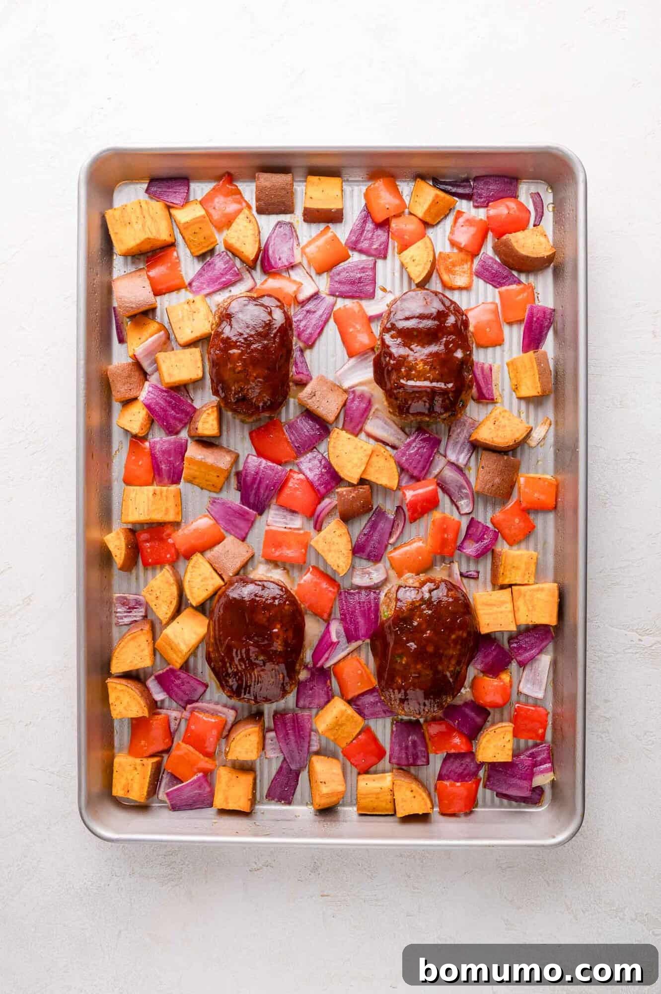 Sheet Pan BBQ Mini Meatloaves 12 Sheet pan with four glazed bbq mini meatloaves surrounded by diced sweet potatoes, red onions, and red peppers, all evenly spaced on a metal tray.