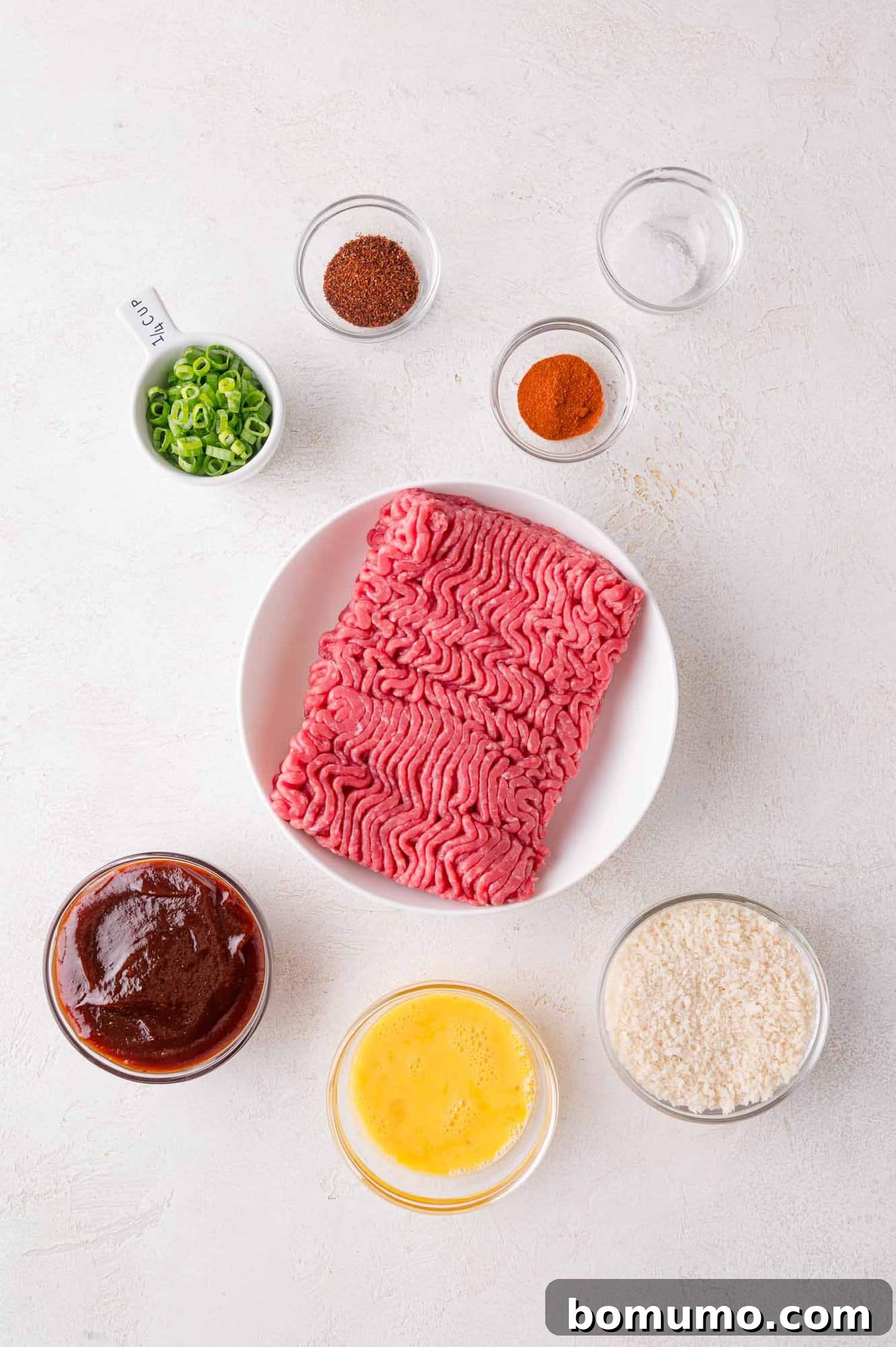 Sheet Pan BBQ Mini Meatloaves 3 Raw ground beef in a white bowl surrounded by small bowls of breadcrumbs, beaten egg, barbecue sauce, sliced green onions, chili powder, paprika, and a clear liquid on a light surface.