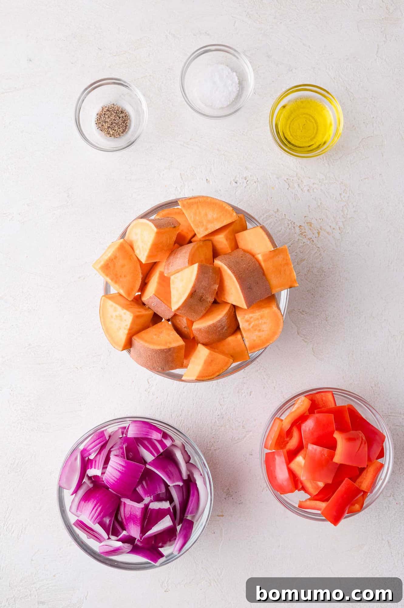 Sheet Pan BBQ Mini Meatloaves 4 Bowls with chopped sweet potatoes, red onion, and red bell pepper, alongside small bowls of salt, pepper, and olive oil, arranged on a white surface.