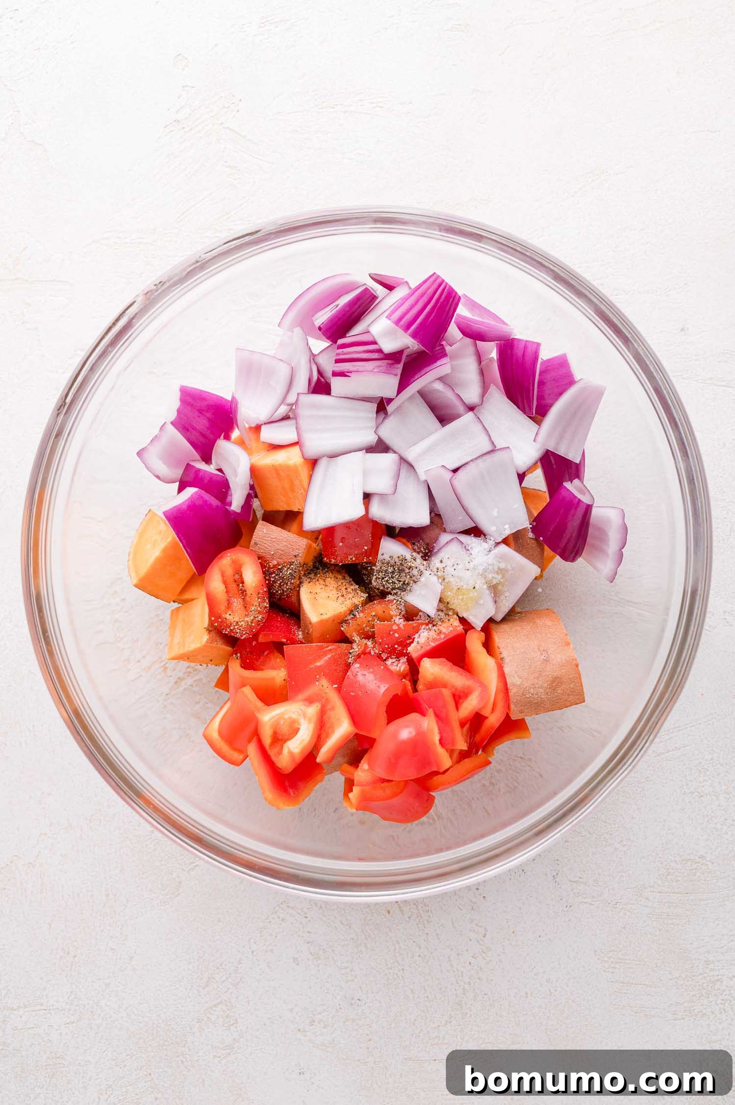 Sheet Pan BBQ Mini Meatloaves 5 A glass bowl containing chopped red onion, sweet potato, red bell pepper, and seasonings on a white surface.