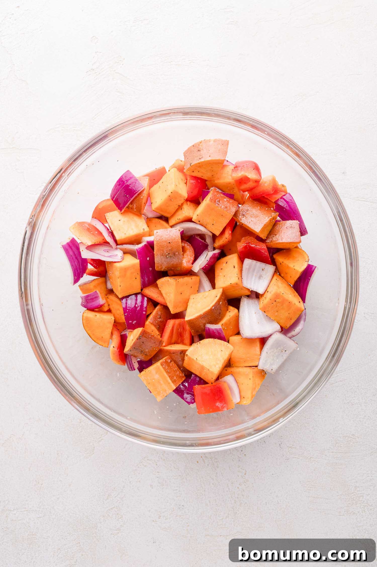 Sheet Pan BBQ Mini Meatloaves 6 A glass bowl containing chopped sweet potatoes, red onions, and red bell peppers, seasoned with spices, on a light surface.