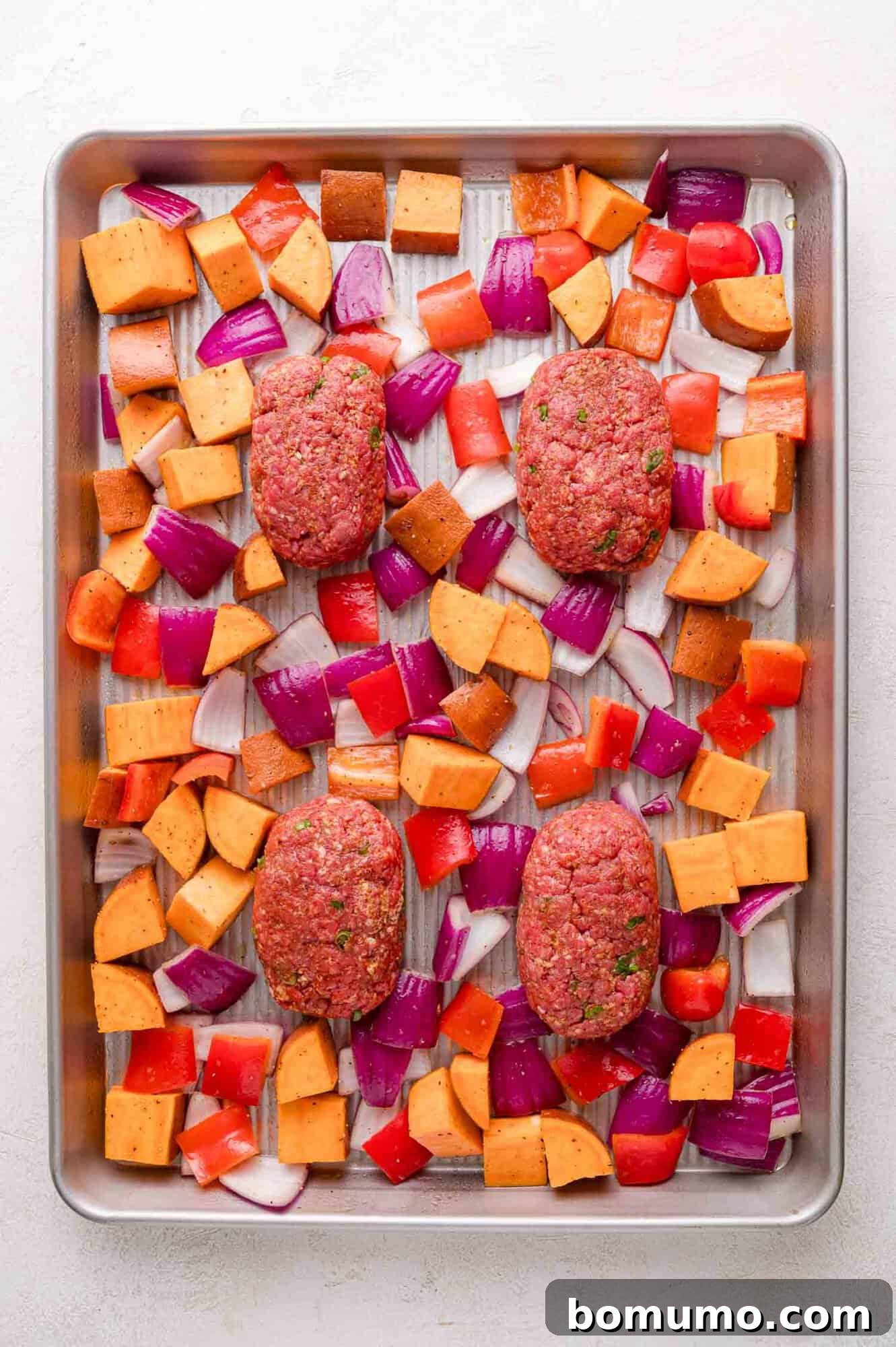Sheet Pan BBQ Mini Meatloaves 10 A baking tray with four raw meatloaf portions, surrounded by chopped sweet potatoes, red onions, and red bell peppers, ready to be cooked.