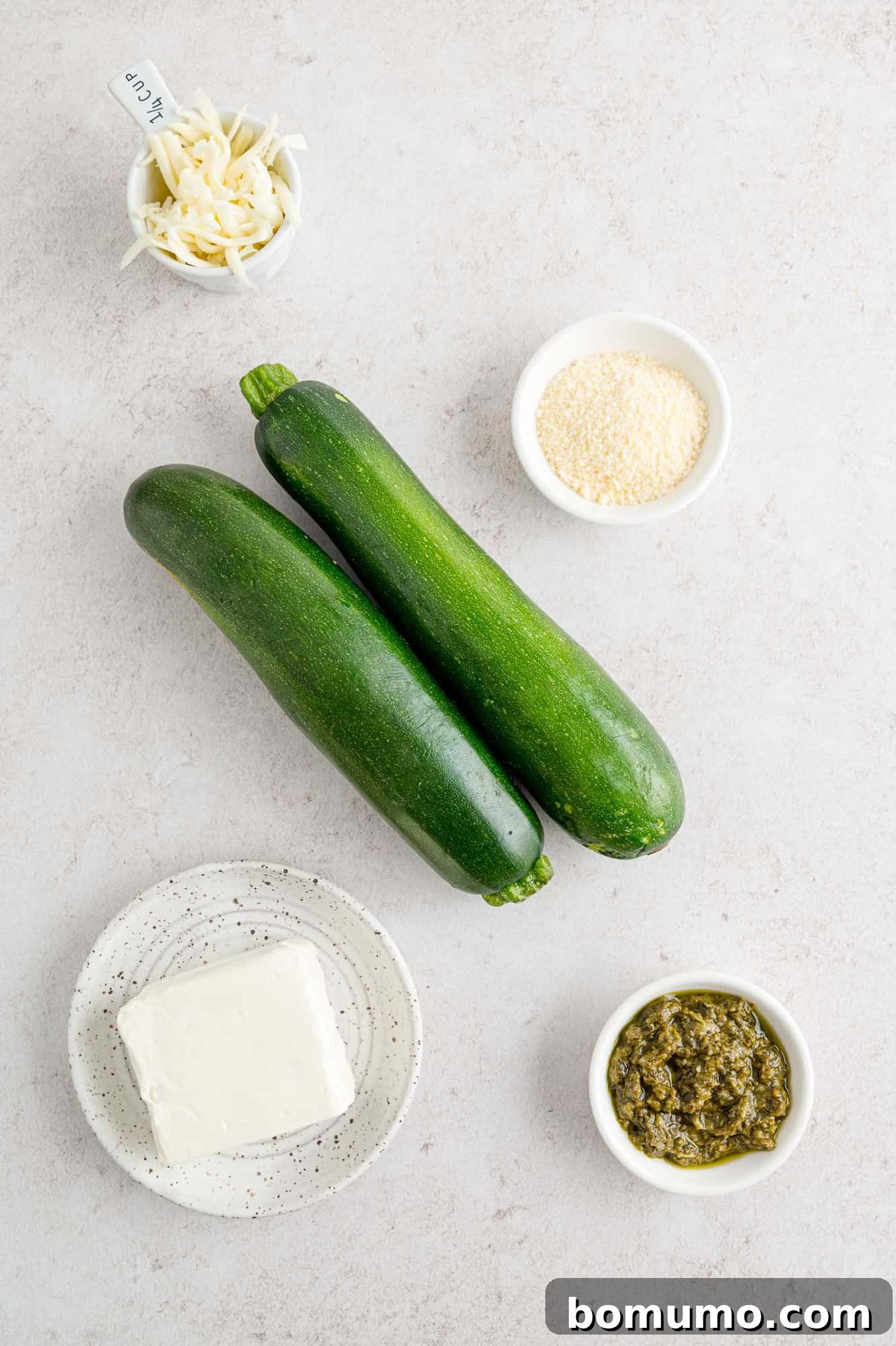 Ingredients for cheese and pesto stuffed zucchini, including two fresh zucchini, various cheeses, and a jar of pesto.