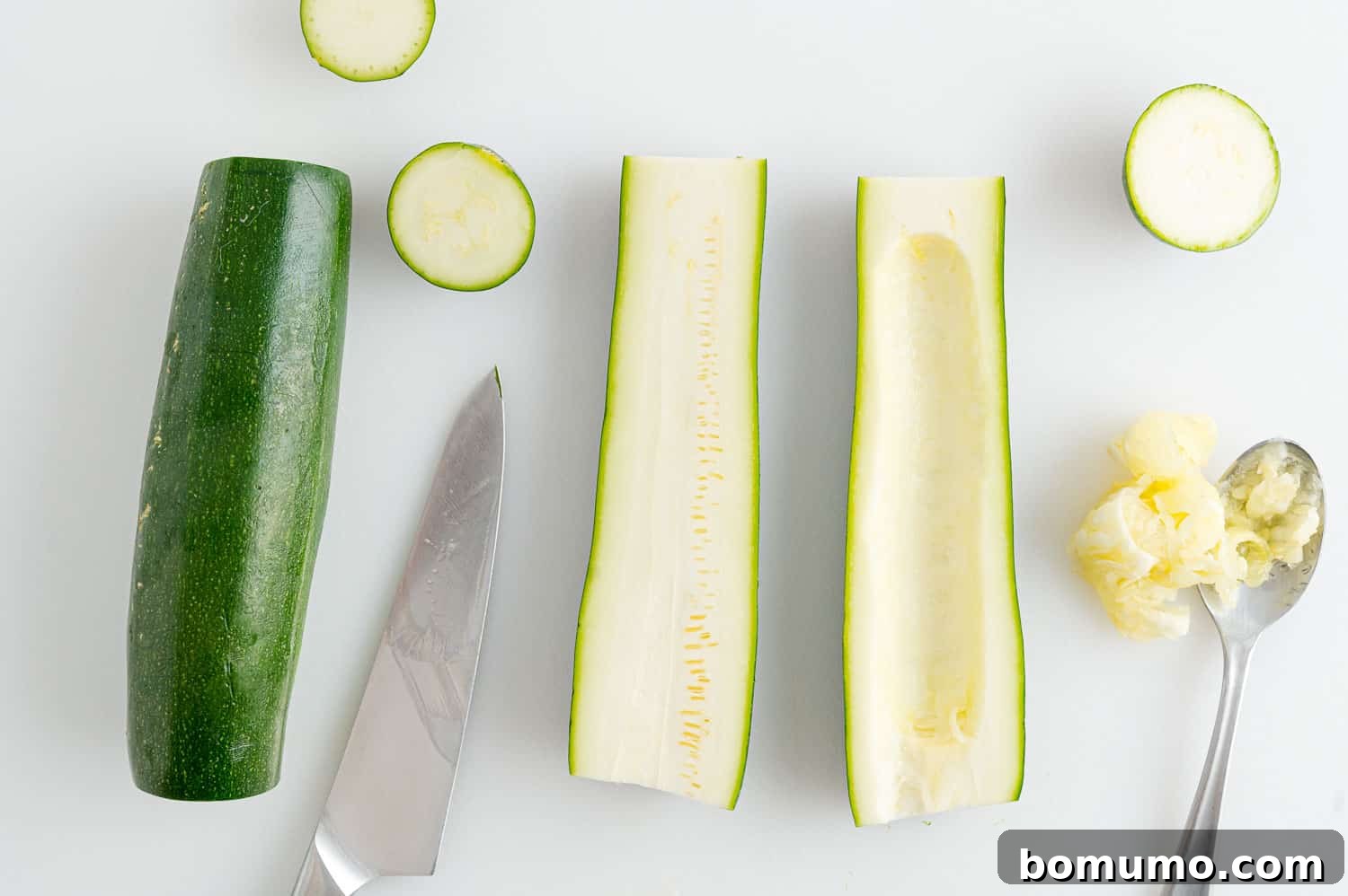 A hand cutting a zucchini in half lengthwise and then scooping out the seeds to create a boat shape.