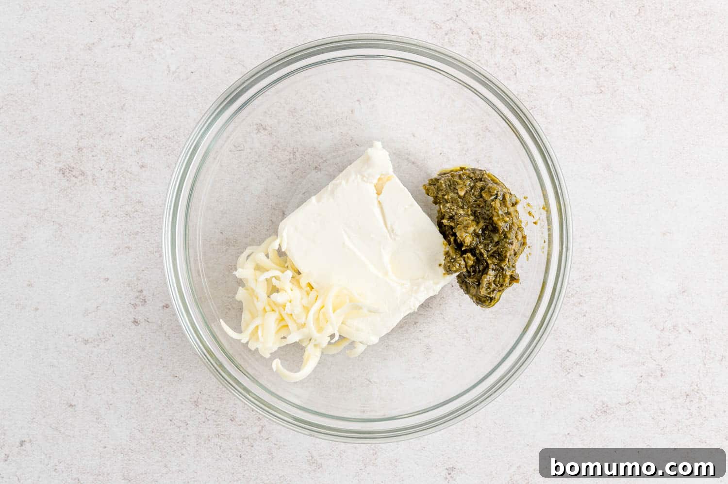 A bowl containing cream cheese, shredded mozzarella, and pesto, before being mixed together.