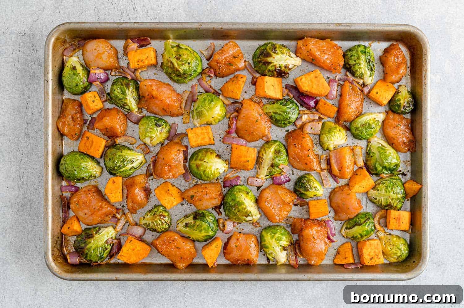 A sheet pan showcasing partially roasted vegetables, including Brussels sprouts, sweet potatoes, and red onion, now joined by raw, seasoned chicken pieces, all arranged in a single layer before the final roasting stage.