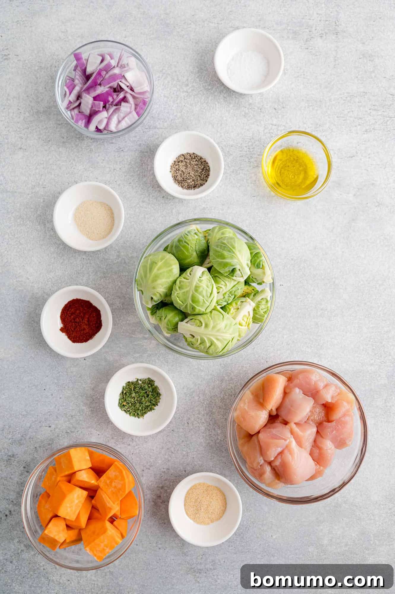 A perfectly arranged display of fresh ingredients for the Hot Honey Chicken Sheet Pan Dinner, including raw boneless chicken breasts, vibrant green Brussels sprouts, a large sweet potato, a red onion, and small bowls containing various ground spices, all ready for meal preparation.