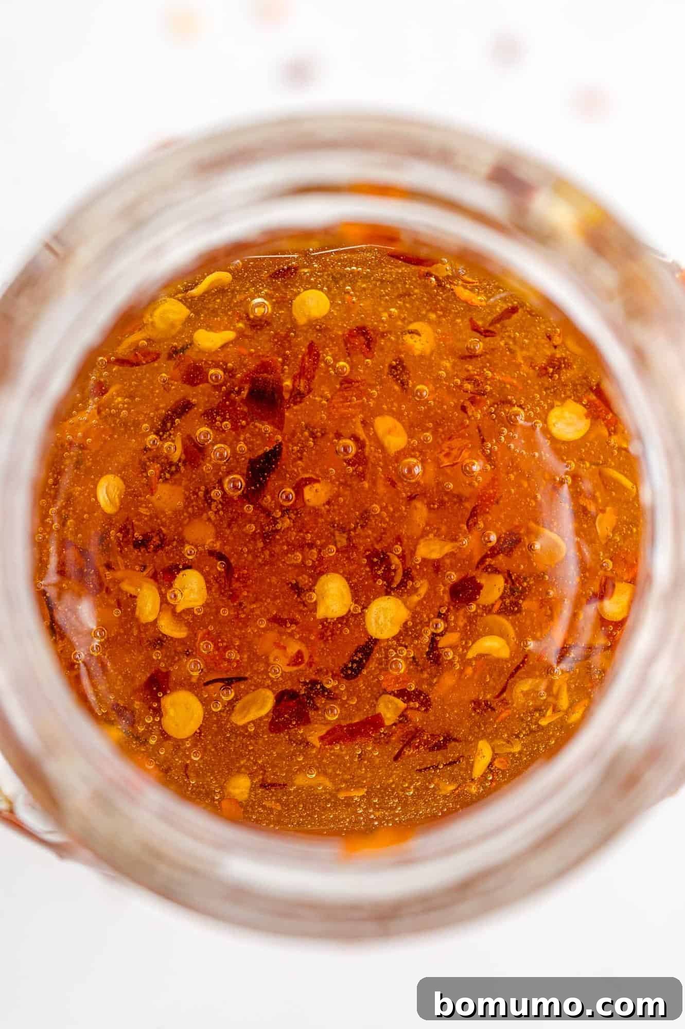 An overhead view of a clear glass jar filled with golden hot honey, clearly showcasing the vibrant red pepper flakes suspended within, symbolizing its sweet and spicy essence.