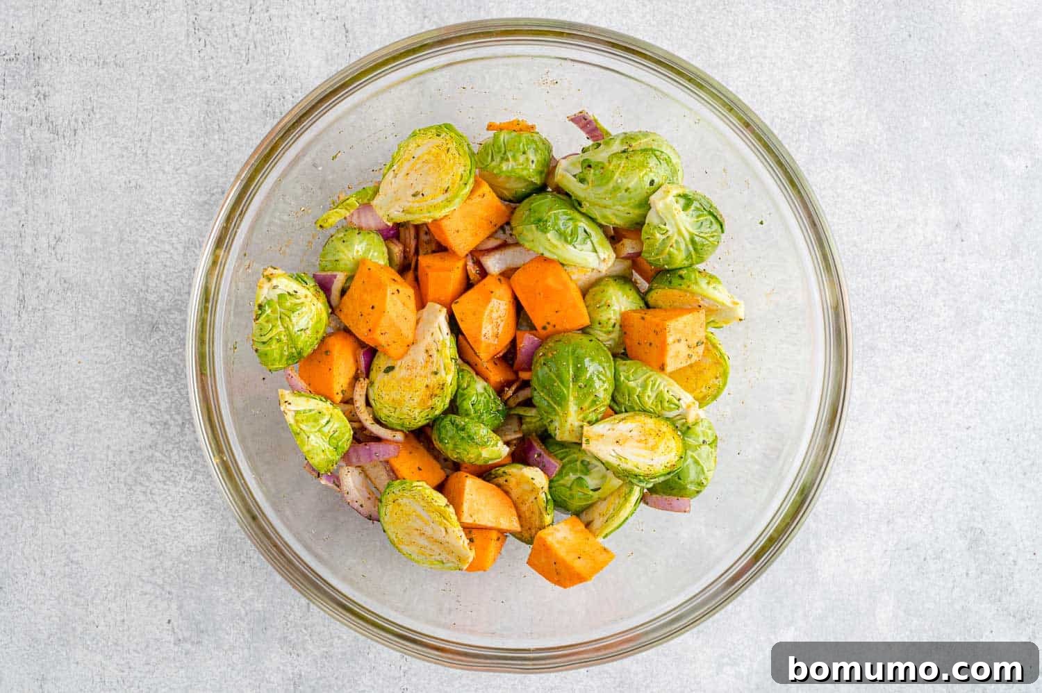 A colorful medley of bite-sized vegetables, including halved Brussels sprouts, cubed sweet potatoes, and chopped red onion, all uniformly coated in a shimmering layer of olive oil and aromatic spices, prepared for roasting.
