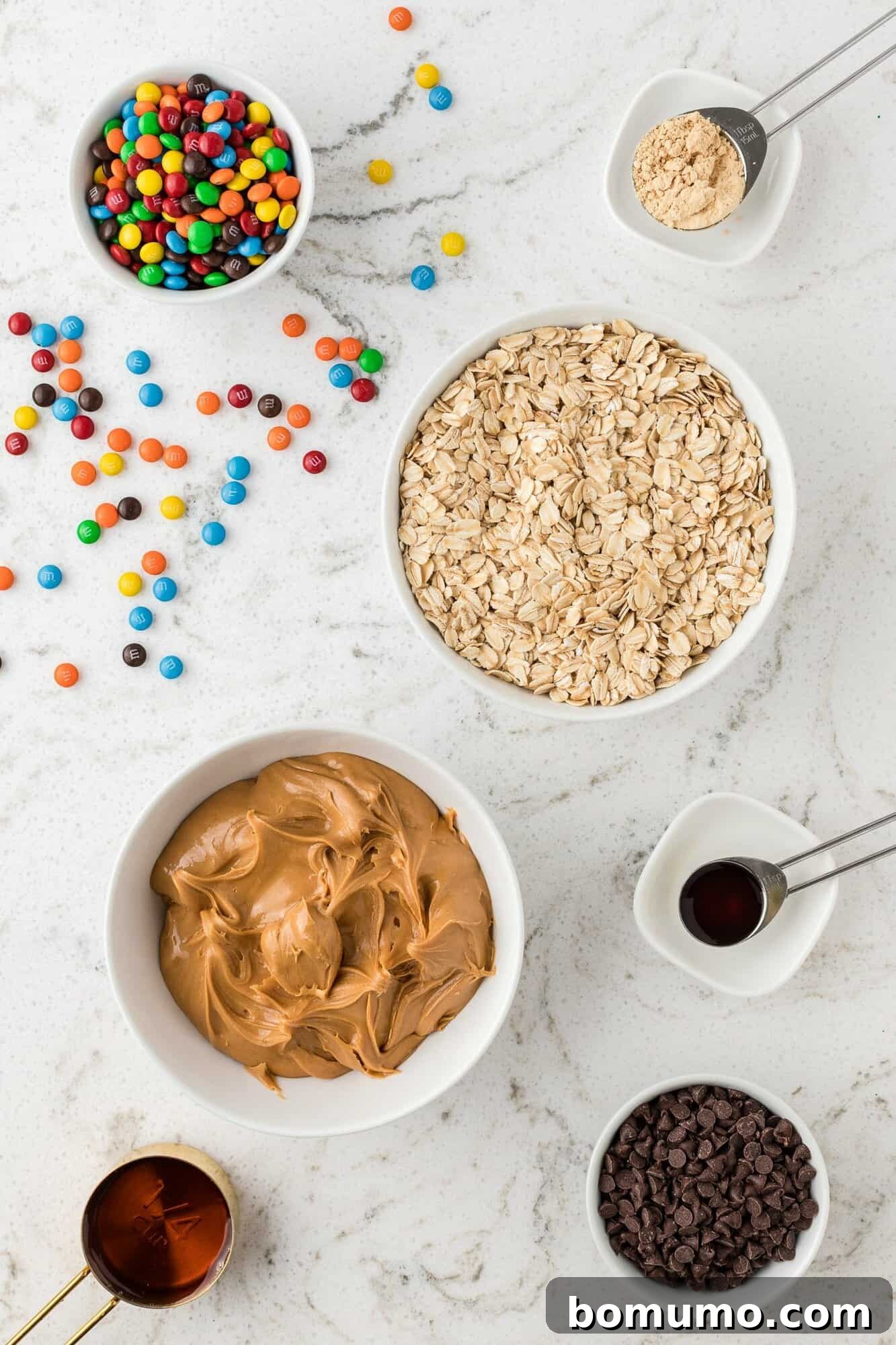 An overhead shot showcasing the key ingredients for Monster Cookie Energy Balls, including creamy peanut butter, wholesome rolled oats, vibrant M&M's, and decadent chocolate chips.