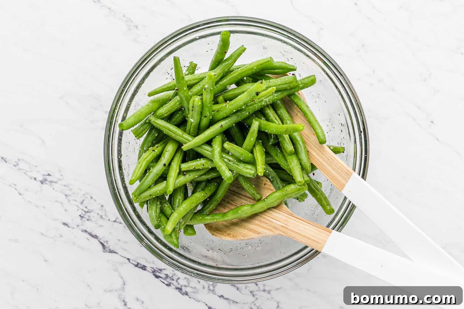 Green beans actively being tossed in a bowl with olive oil and spices, ensuring even coating.