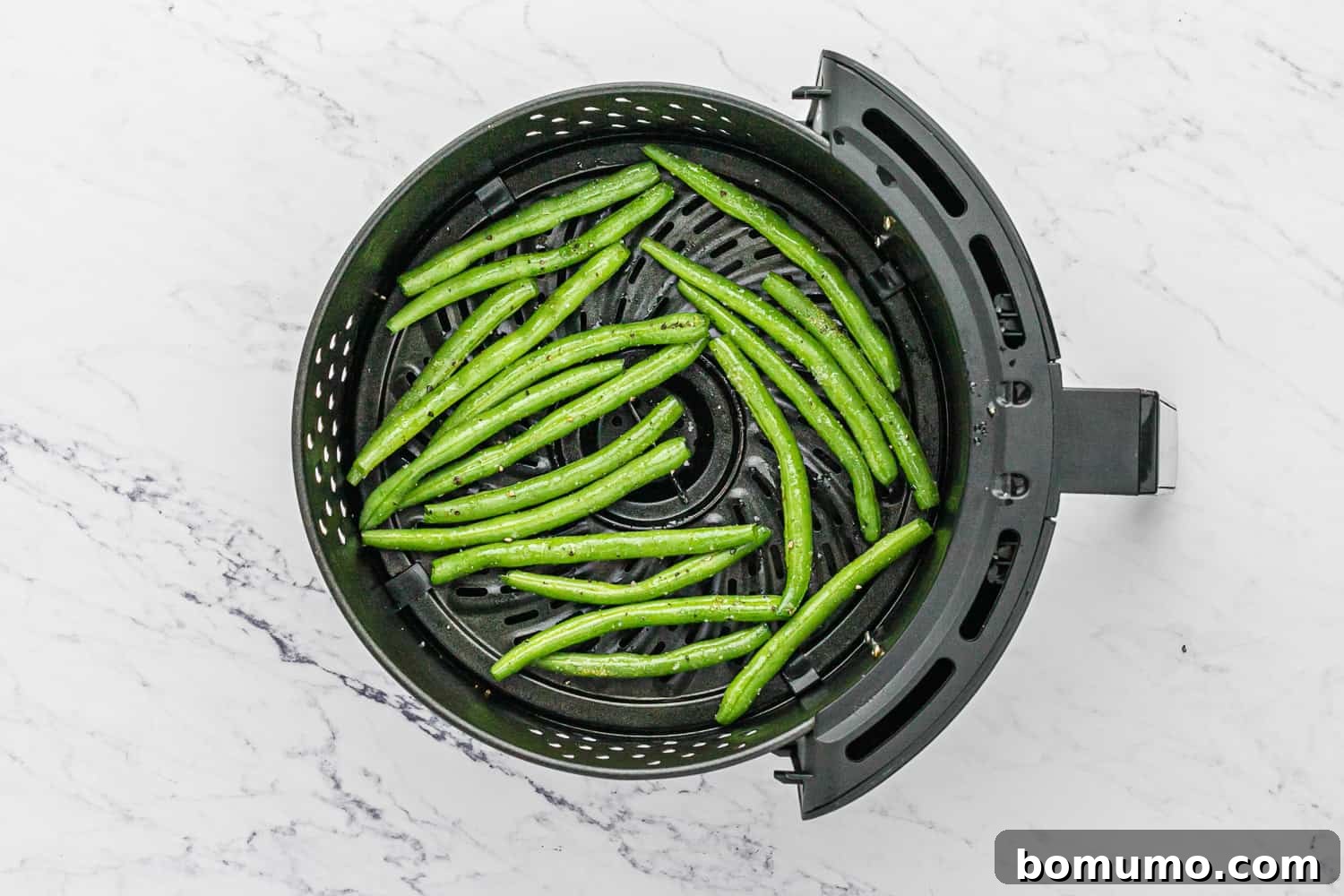 Uncooked, seasoned green beans spread out in a single layer within an air fryer basket, ready for cooking.
