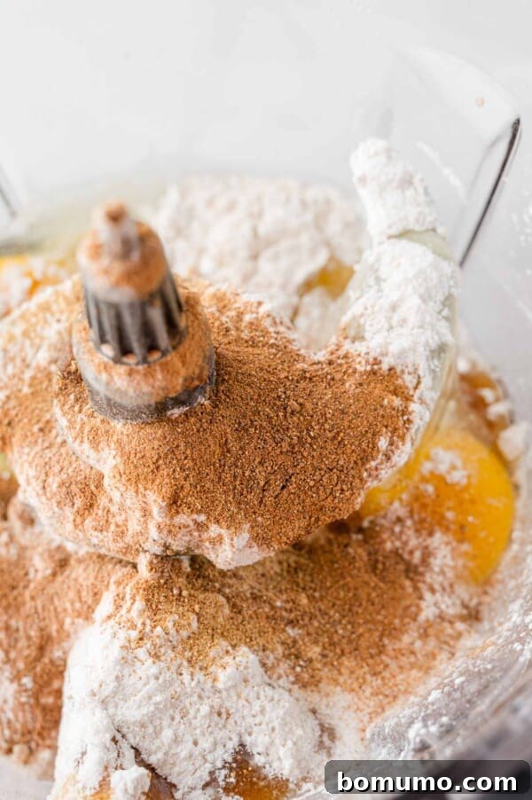 The vibrant orange of cooked carrots is visible inside the bowl of a food processor, ready to be blended into a smooth soufflé batter.