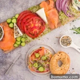 Bagel with smoked salmon, cucumber, tomato, onion, alfalfa sprouts, and cream cheese on a plate, next to a wooden board with more toppings, on a gray surface.