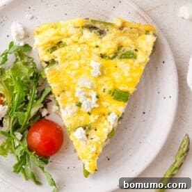A vibrant slice of vegetable frittata with prominent asparagus and goat cheese, served on a plate with fresh mixed greens and a cherry tomato.