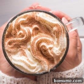 A person holding a glass mug of whipped cream-topped cinnamon latte, sprinkled with cinnamon, on a cozy background.