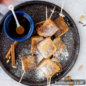 French toast skewers sprinkled with powdered sugar, with a small bowl of maple syrup on the side.