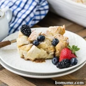A slice of bread pudding with scattered blackberries and blueberries, alongside a fresh strawberry, on a white plate.