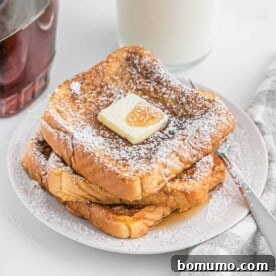 French toast stacked three high on a white plate, elegantly topped with a pat of melting butter and a dusting of powdered sugar.