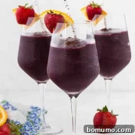 Three long stem wine glasses filled with frozen sangria, and topped with a fresh strawberry and orange slice. A paper straw is in each glass.