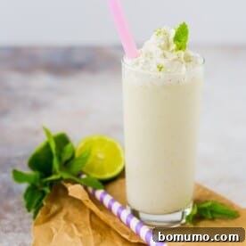 A tall glass of mojito milkshake topped with whipped cream and a mint leaf, with a pink straw. Beside it are a lime half, mint sprigs, and a purple-striped straw.