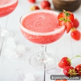 Frosé in a glass, garnished with a strawberry slice, surrounded by whole strawberries and ice cubes on a white surface.