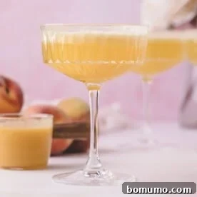 A coupe glass filled with a light orange cocktail, with peaches and another glass in the background on a light pink surface.
