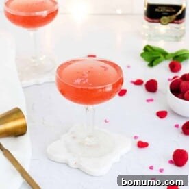 Light red drink in a stemmed glass with raspberries nearby.