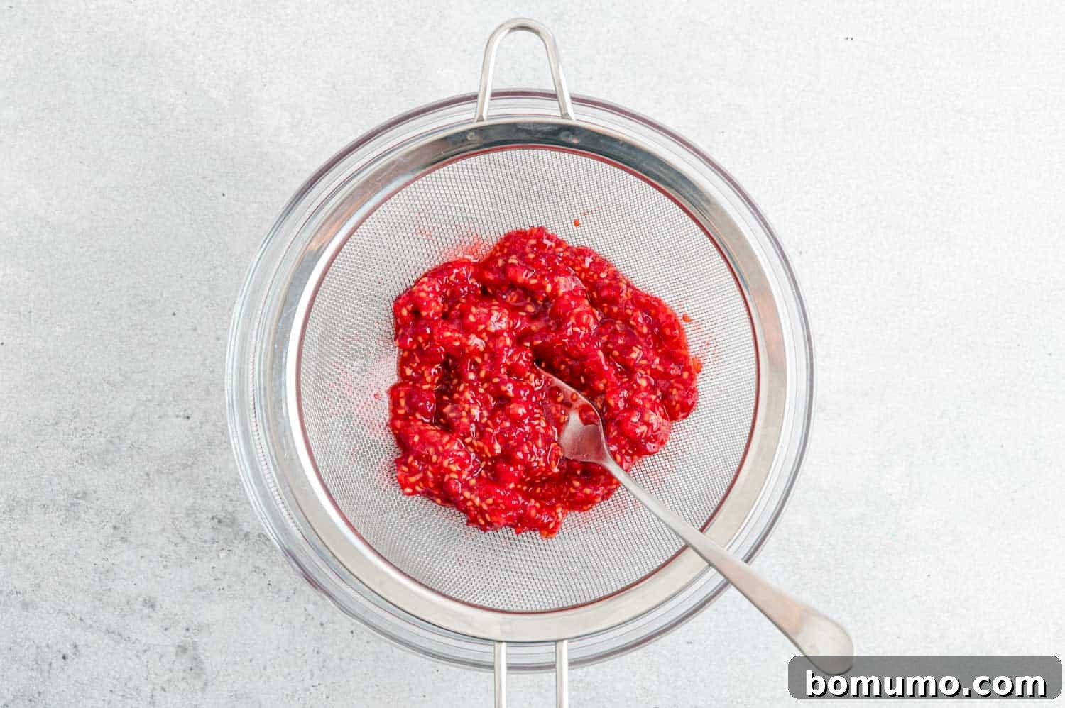 Fresh raspberries being mashed with a fork in a fine mesh strainer over a bowl, illustrating the first step of puree preparation.