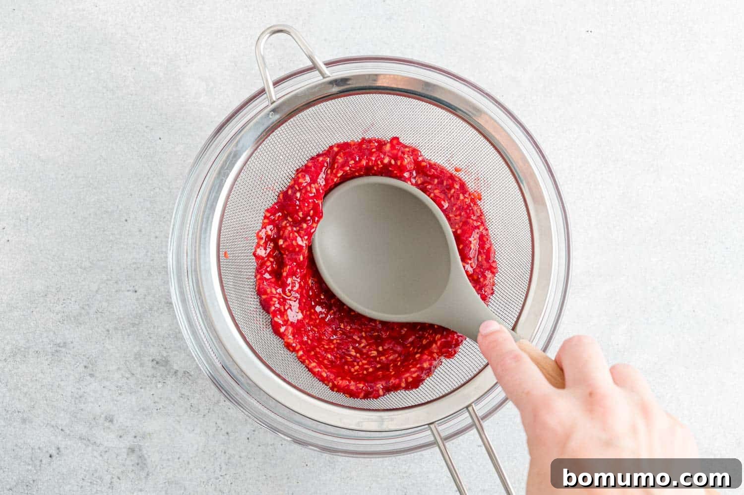 A spoon skillfully pressing raspberry puree through a fine mesh strainer, ensuring a smooth, seedless base for the vinaigrette.