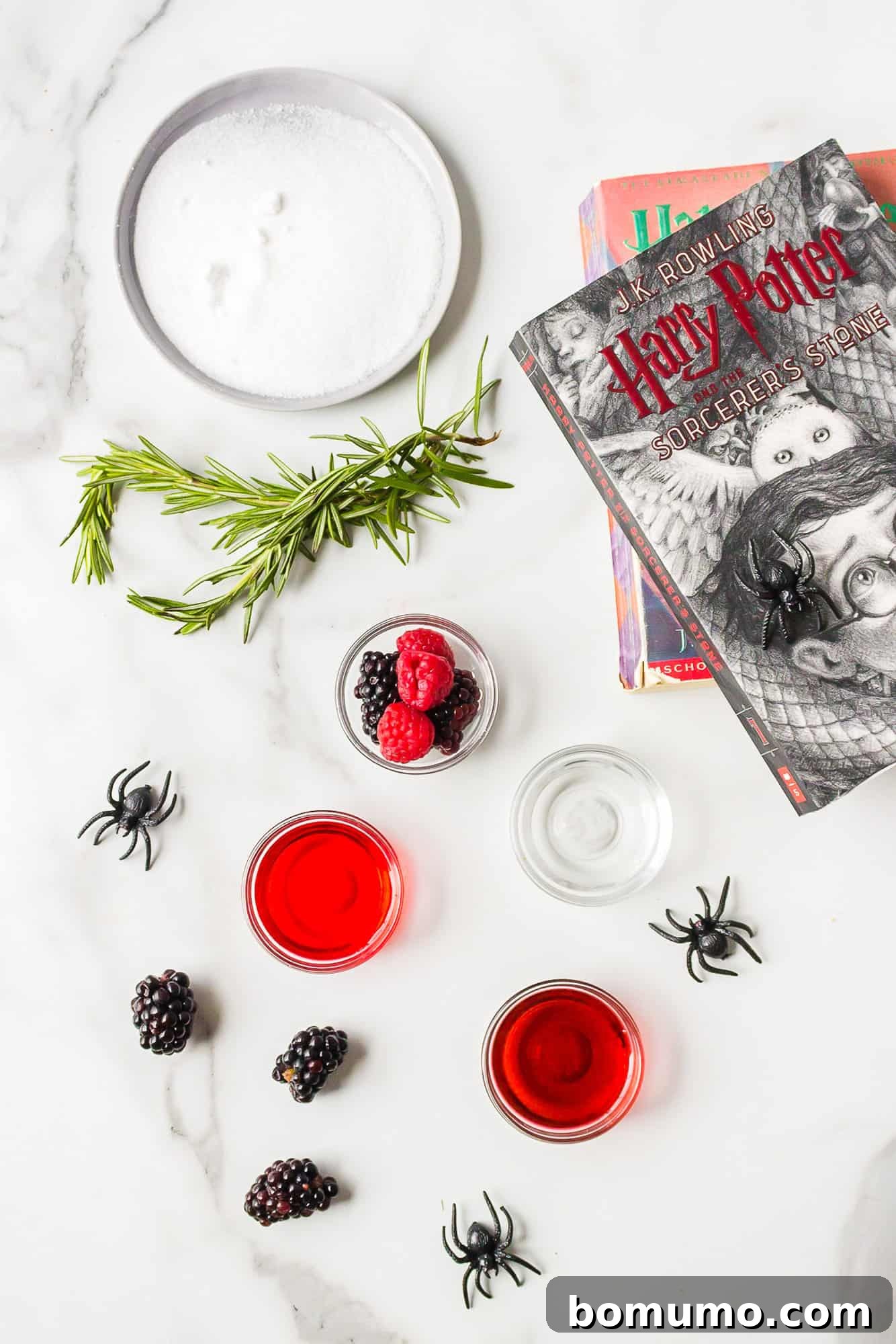 All the ingredients laid out for the Love Potion Cocktail, including raspberry liqueur, vodka, grenadine, sparkling water, and fresh berries, with playful fake spiders scattered around for a Halloween twist.