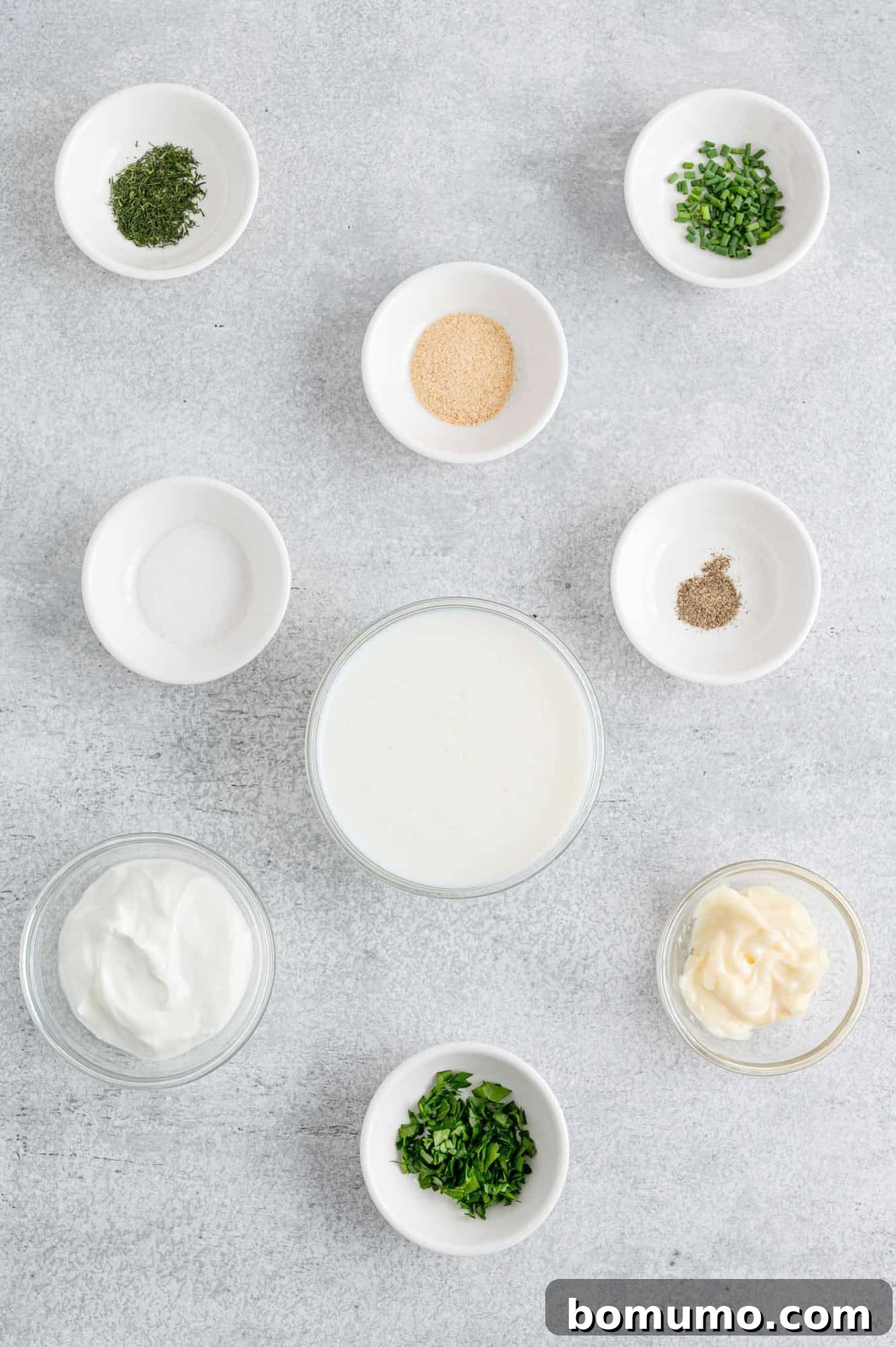 Individual bowls showcasing the fresh and dried ingredients needed for healthy ranch dressing, including vibrant green herbs.
